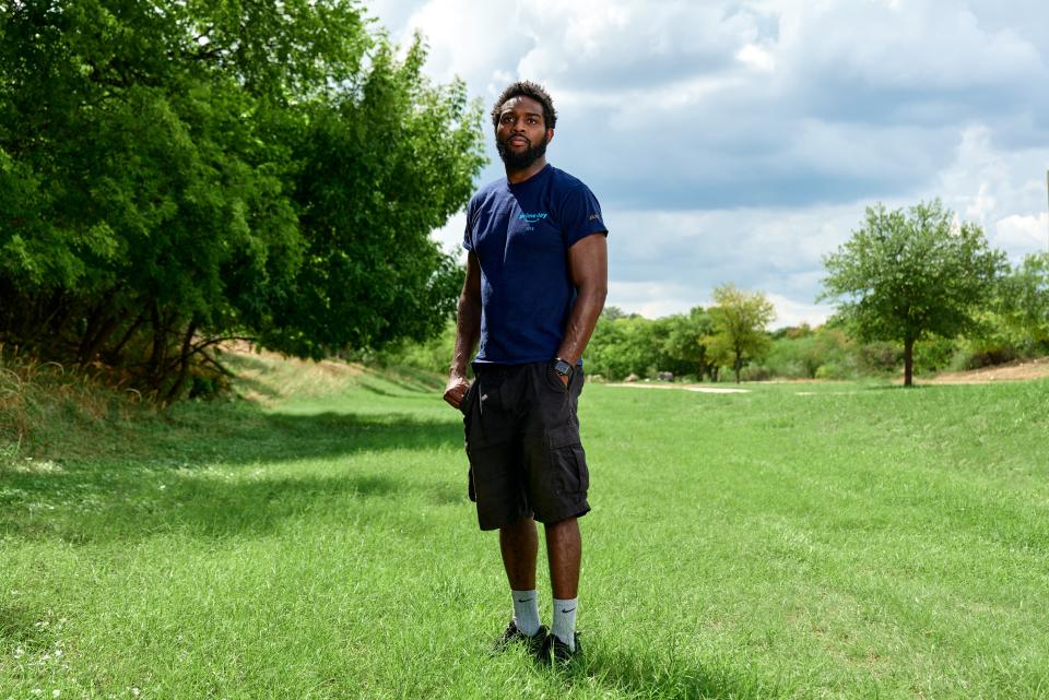 Warehouse worker Daniel Olayiwola stands on a green lawn near trees, in his Amazon uniform.