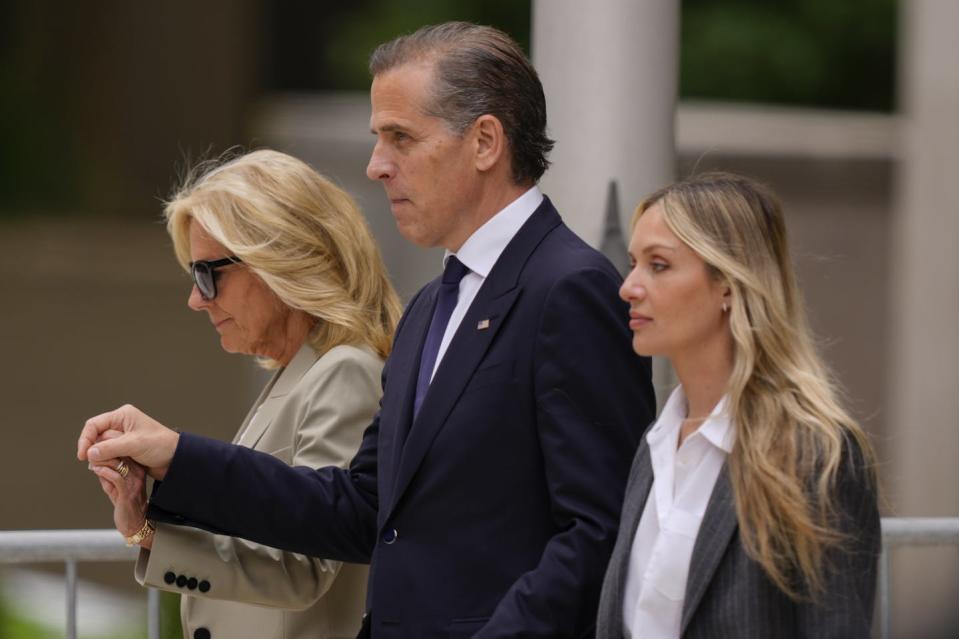 A middle-aged man wearing a dark suit holds hands with a woman with blonde hair, and walks next to another woman, also blonde and younger.