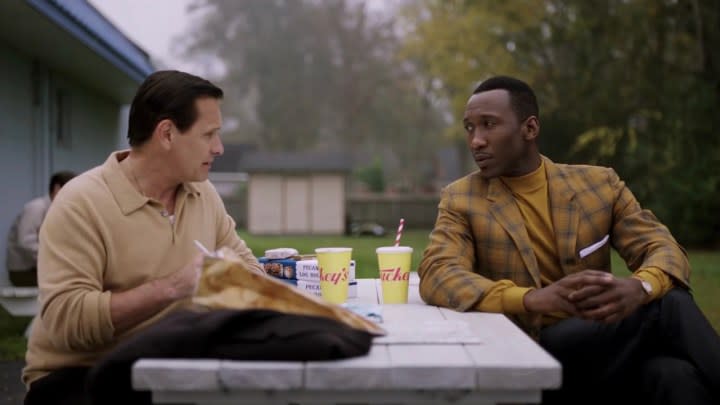 Two men sit at a picnic table outside eating and talking in a scene from Green Book.
