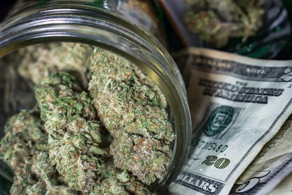 Cannabis buds in a glass jar lying atop a small stack of cash.