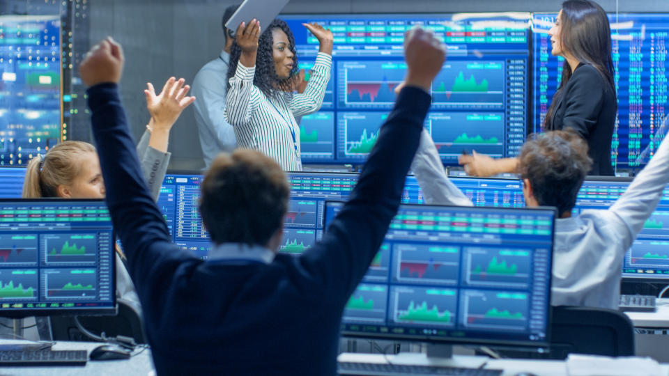 Traders celebrating in front of colorful monitors displaying stock market data.