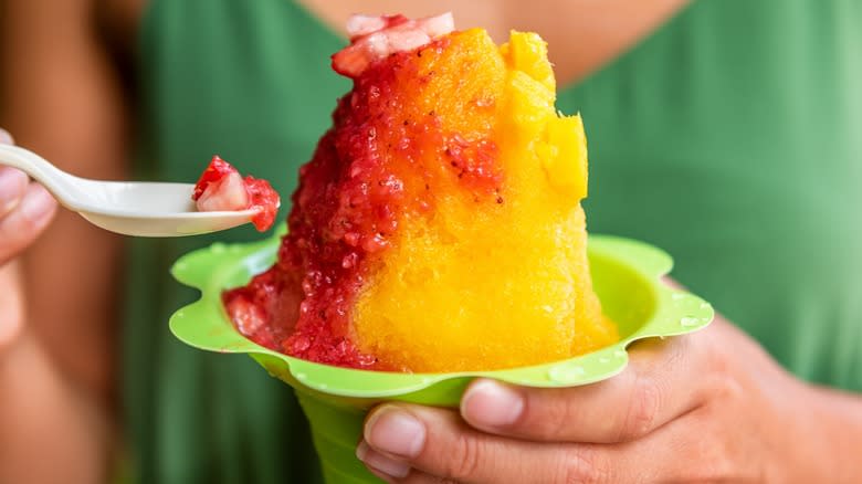Hand holding a yellow and red Hawaiian shaved ice and a plastic spoon