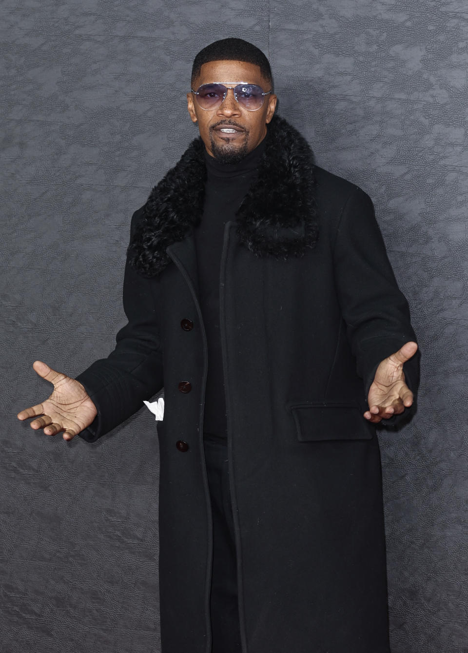 Jamie Foxx poses in a stylish black coat with fur trim and purple-tinted sunglasses against a plain background