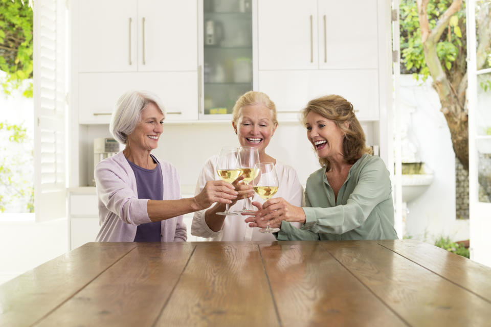 Three women, smiling and clinking glasses, seated at a wooden table in a bright kitchen