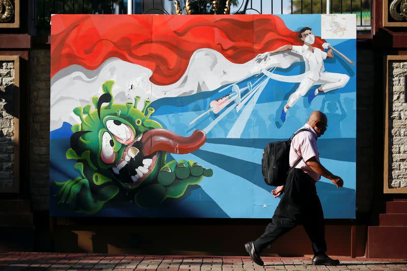 A man walks past a mural depicting a character attempting to kill the coronavirus with the vaccine, in Jakarta
