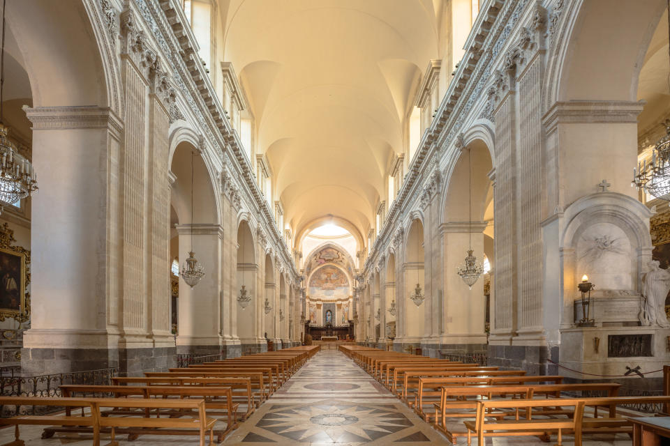 Interior of a grand, empty cathedral with high arches, ornate decorations, and wooden pews lined up along the marble floor leading to an altar at the back