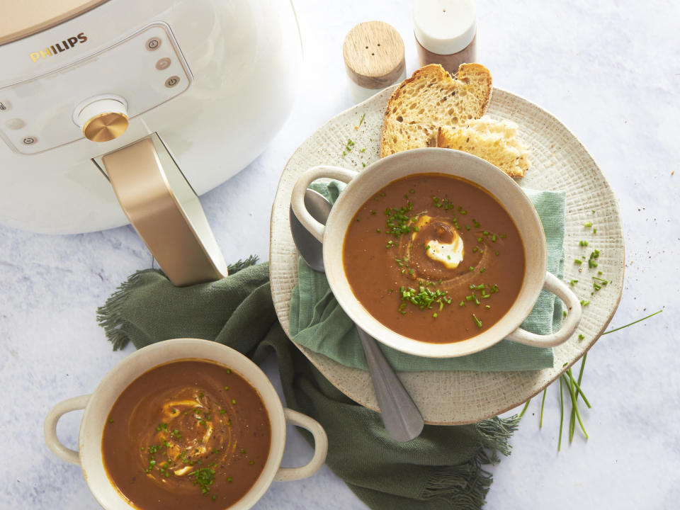 A white bowl with handles holding honey roasted pumpkin soup and a swirl of sour cream, sprinkled with chives sitting on a green napkin and beige plate with broken toast beside. Seen from above, another bowl of soup sits to one side and a white and gold Philips air fryer can be seen.