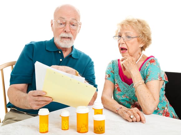 A visibly worried senior couple looking at their medical bills, with an assortment of prescription medicine bottles in front of them.