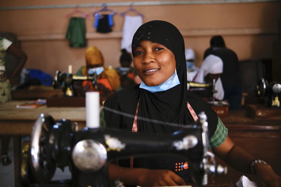 A young woman trainee sits at a sewing machine wearing a black head covering, a Covid mask pulled beneath her chin, a tape measure around her neck and is smiling for the camera.