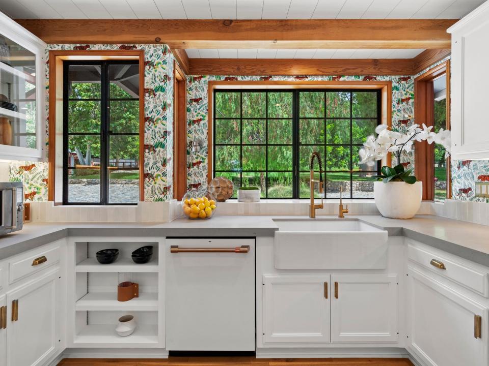 A white kitchen counter with multiple drawers and cabinets, a large window looking out at a tree, and white flowers