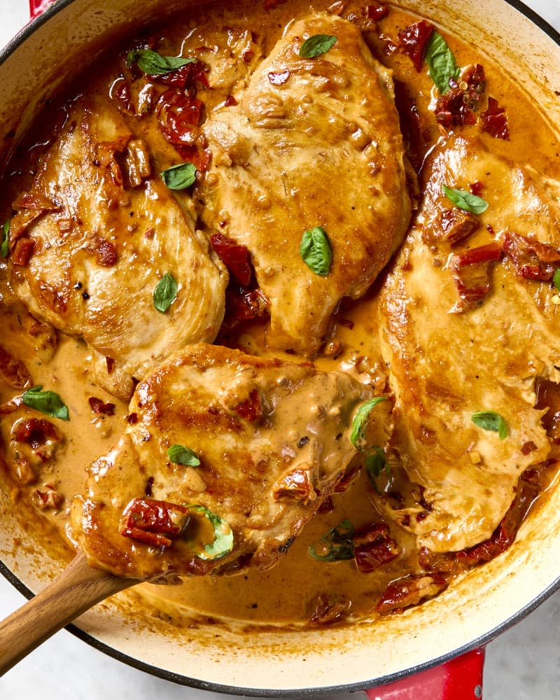 overhead shot of four pieces of marry me chicken in a red dutch oven, with a wooden spoon lifting a piece of chicken out of the pot.