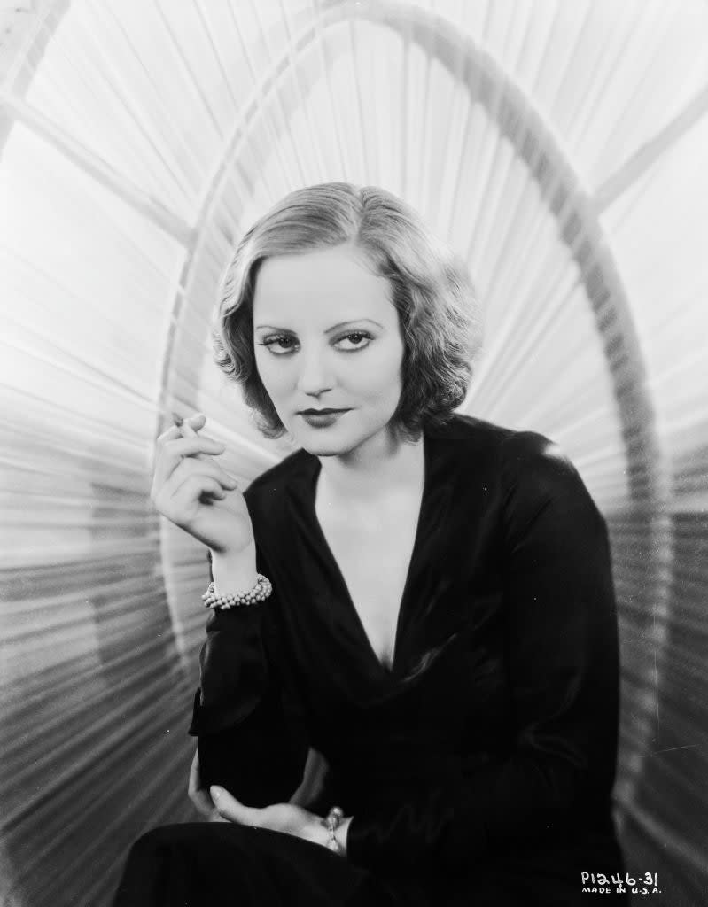 A black-and-white image of a woman in a satin dress with a deep neckline, holding a cigarette