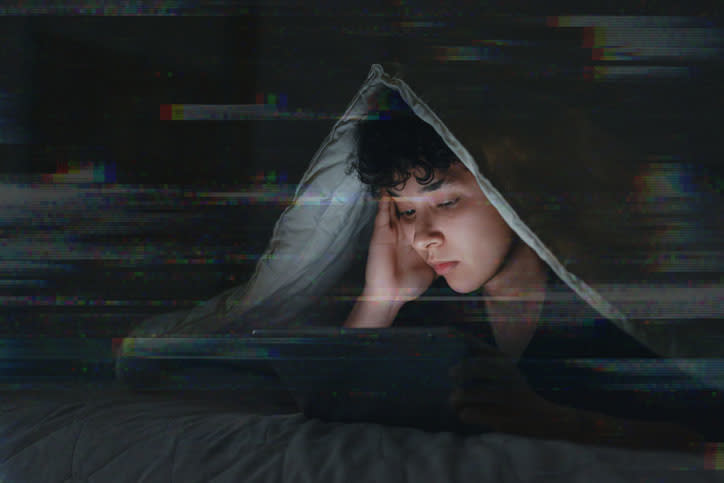 Person under a blanket looks at laptop screen in a dimly lit room