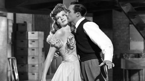 rita hayworth stands next to gene kelly who wraps one arm around her waist and kisses her cheek, she wears a ball gown, he wears slacks, a white collared shirt, and a dark sleeveless sweater vest
