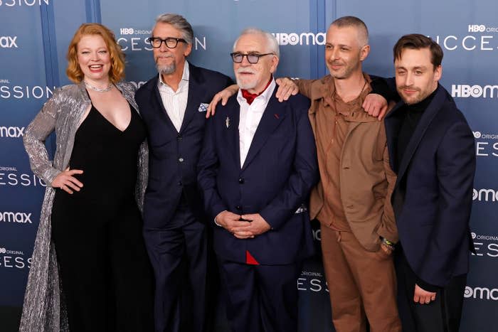 Five cast members from "Succession" posing; one in a silver gown, three in suits, and one in a brown ensemble