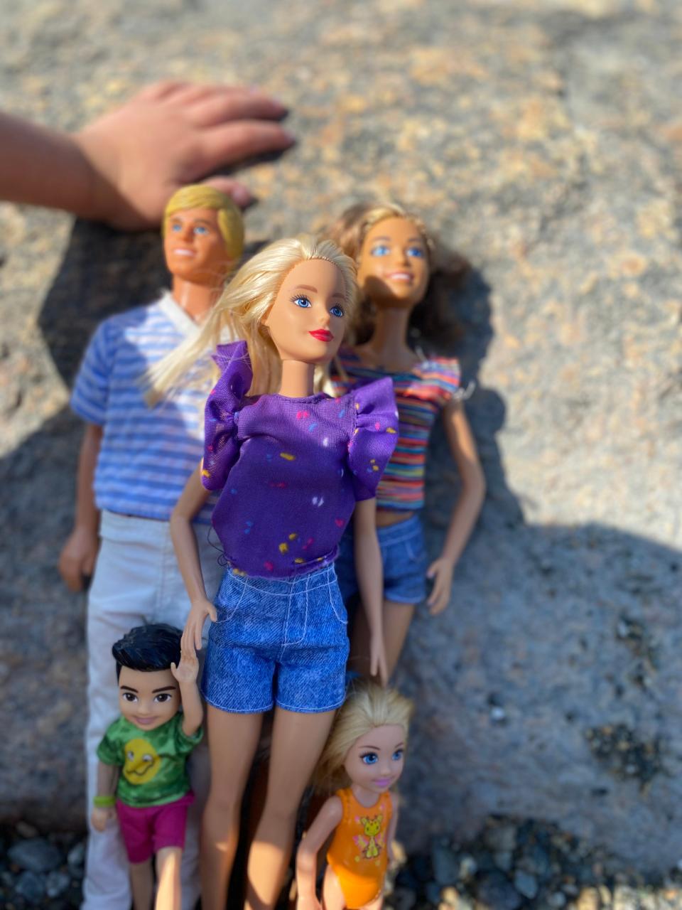 Image of 5 dolls laying on a rock. The doll on the left is a Ken doll with short blonde hair and blue eyes, wearing a blue and white striped t-shirt and white pants. The Barbie on the right has light brown hair, blue eyes, and wears a multi-color stripe shirt and jean shorts. On top of both of them lies a blonde Barbie with blue eyes and red lips, wearing s purple shirt with flouncy sleeves and a multi-color dotted pattern, along with jean shorts. On her left and right are two baby Barbie dolls. The one on the left is a boy with dark brown hair and dark eyes, with a neon green shirt that has a yellow logo on it and pink shorts. The other baby doll is a girl with long blonde hair and an orange dress that has a cartoon giraffe. To the left of the dolls a toddler's hand rests on the rock and the toddler's shadow falls on the dolls. The rest of the rock is in the sun. 