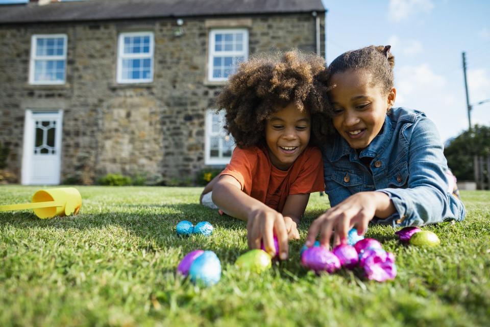 two siblings lying down in the grass on their front smiling while holding their easter eggs