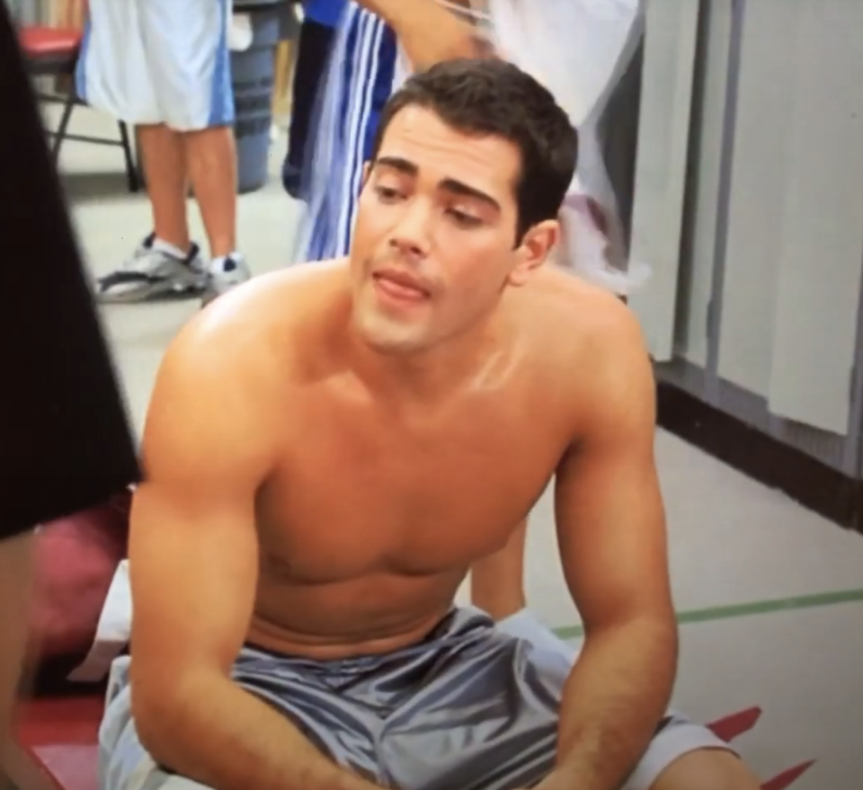 Jesse Metcalfe is sitting shirtless in a gym, resting with his tongue slightly out. Other people are seen in the background