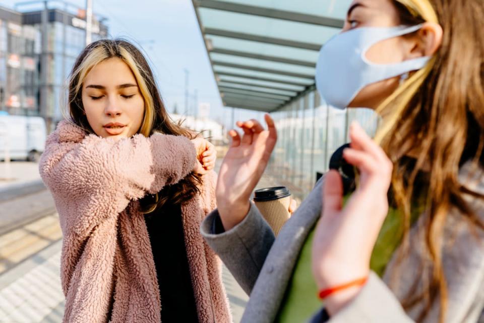 Young sick student teenager woman outside at bus stop is sneezing into the elbow from an allergy or cold.  Scared woman in protective mask afraid to cough woman outdoor