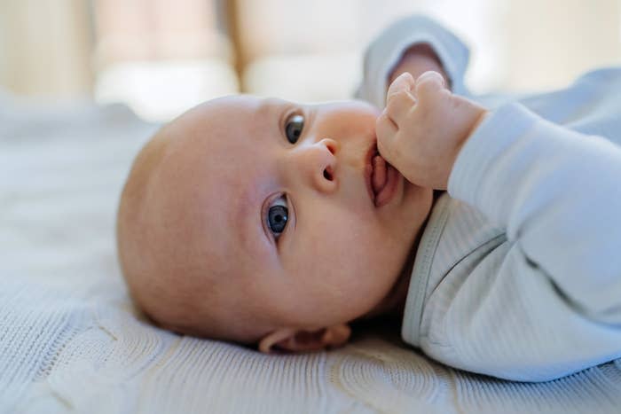 Baby lying on back, sucking thumb, wearing a onesie, looking thoughtfully