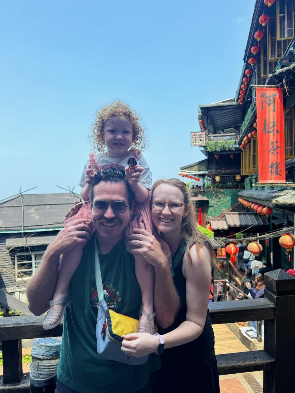 A couple poses in front of a marketplace. Their daughter sits on her father's shoulders.