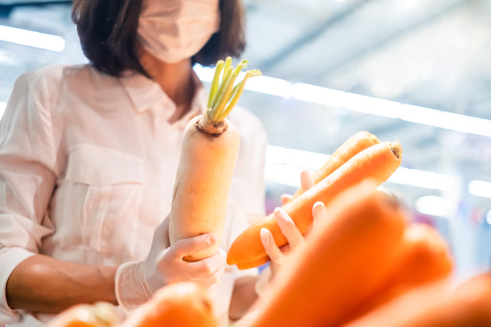 Asian woman wearing protect face mask and rubber gloves shopping fruit, vegetable in grocery department store. Girl choosing carrot, radish in supermarket during coronavirus crisis, covid19 outbreak.