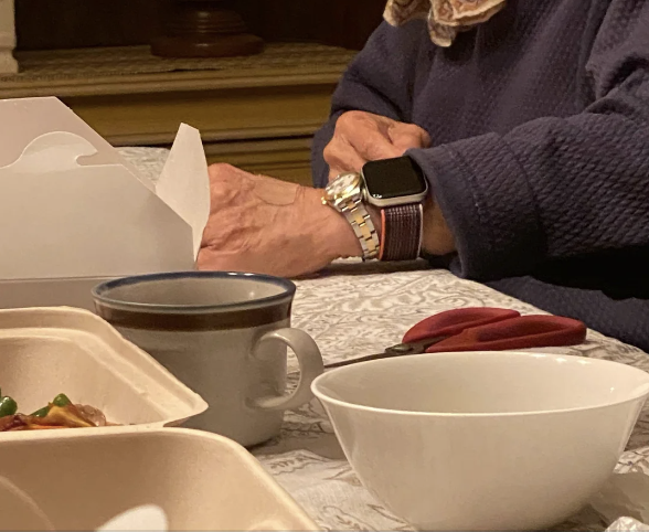 An elderly person's hands are seen on a table, one hand wearing a smartwatch and a bracelet, a white takeout container, a mug, and food containers are nearby