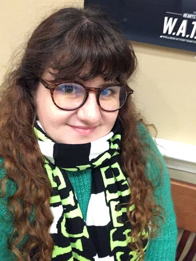 A person with long curly hair, wearing glasses, a green sweater, and a patterned scarf, smiles at the camera