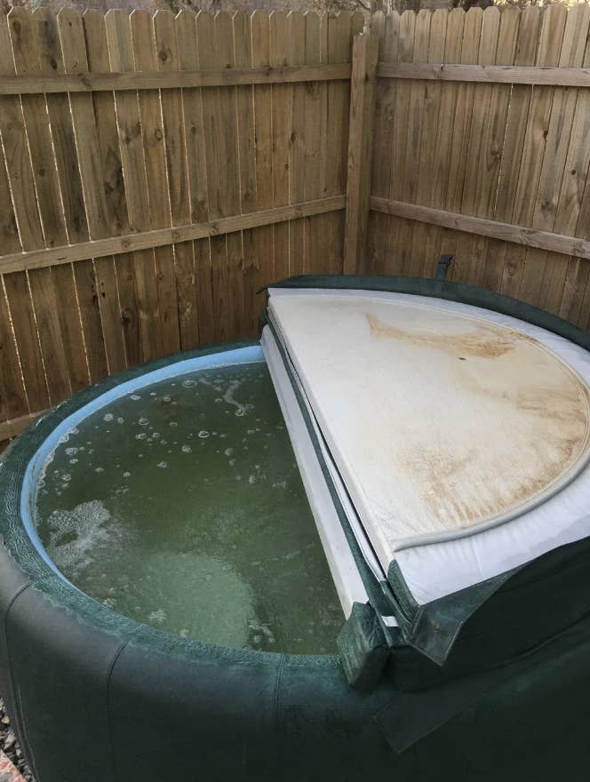 Hot tub with partial cover next to a wooden fence, water appears unclean