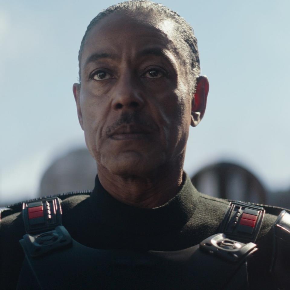 Giancarlo Esposito in a black uniform with red straps, standing with a serious expression, flanked by two figures in armor