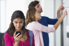 University students checking for test results in bulletin board while woman checking text message on mobile phone