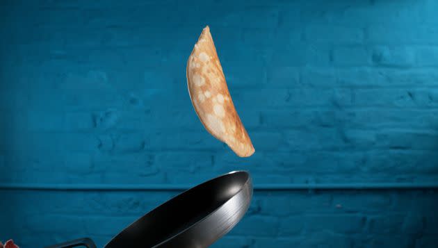 Crepe flipping midair over pan against blue brick wall