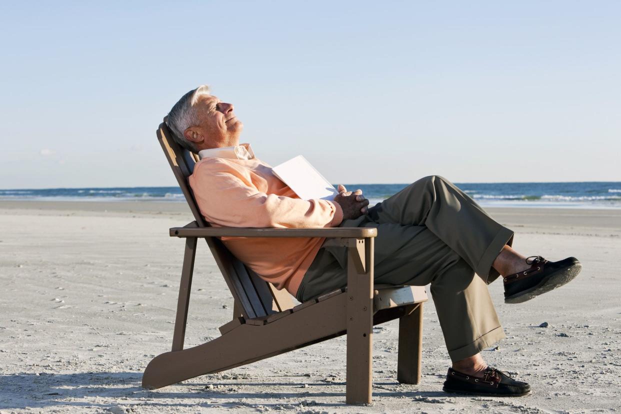 senior man on beach smiling and napping with book in lap, legs crossed