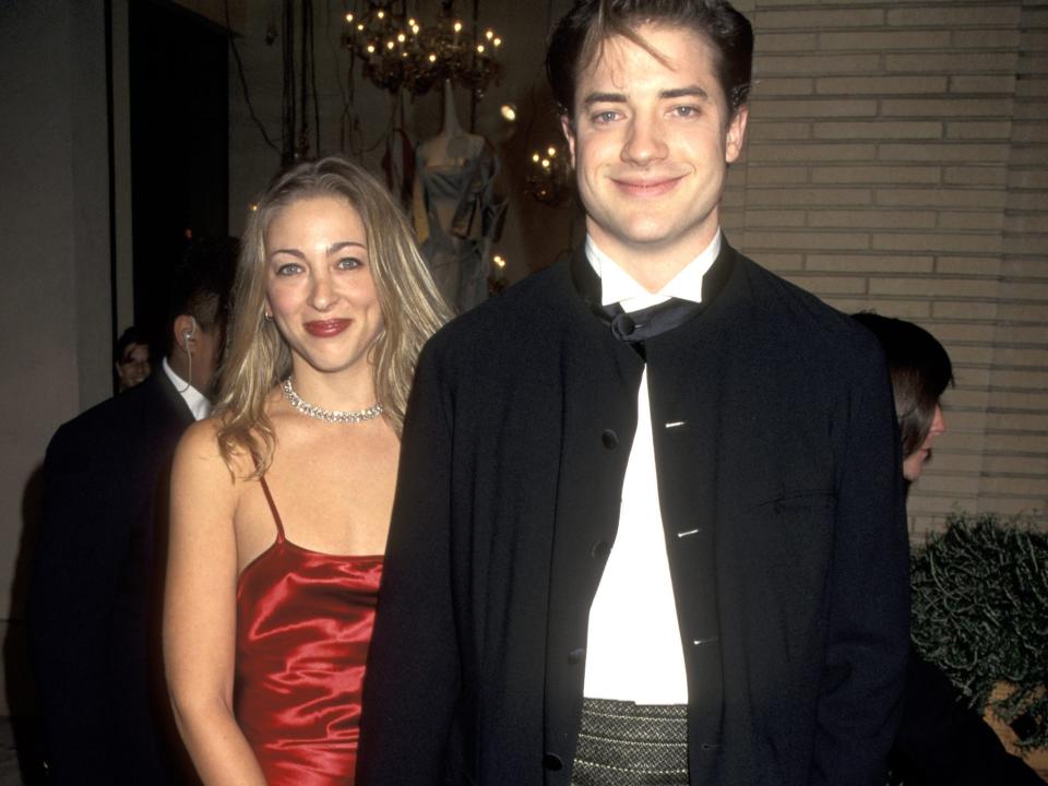 Afton Smith, in a red dress, holds hands with Brendan Fraser, in a black tuxedo, at a 1995 event.