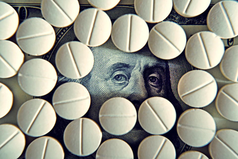 Prescription drug tablets covering up a one hundred-dollar bill, with Ben Franklin's eyes peering out through an opening.