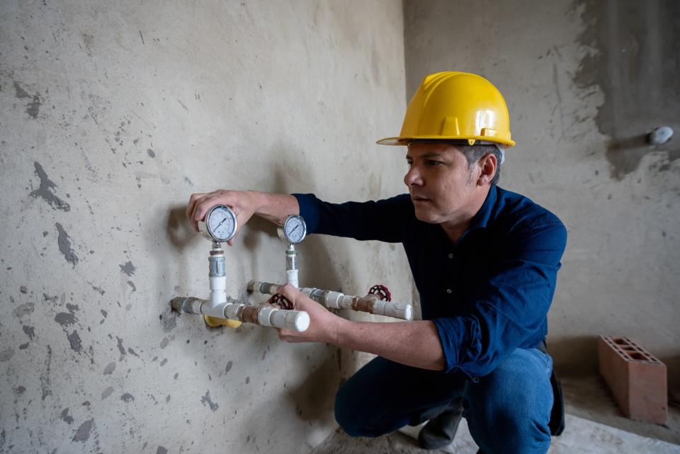 A worker in a yellow hard hat installs two gas lines in a bare wall. 