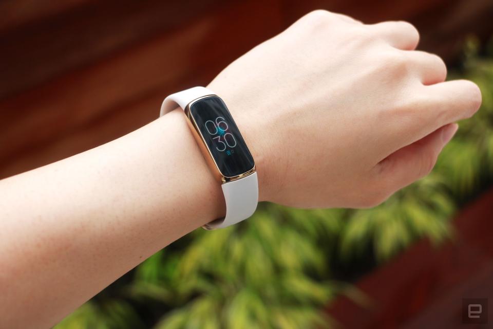 <p>Slight off angle view of the Fitbit Luxe with a light pink silicone band on a wrist against a dark brown background with some greenery. The screen shows the time is 6:30pm.</p> 