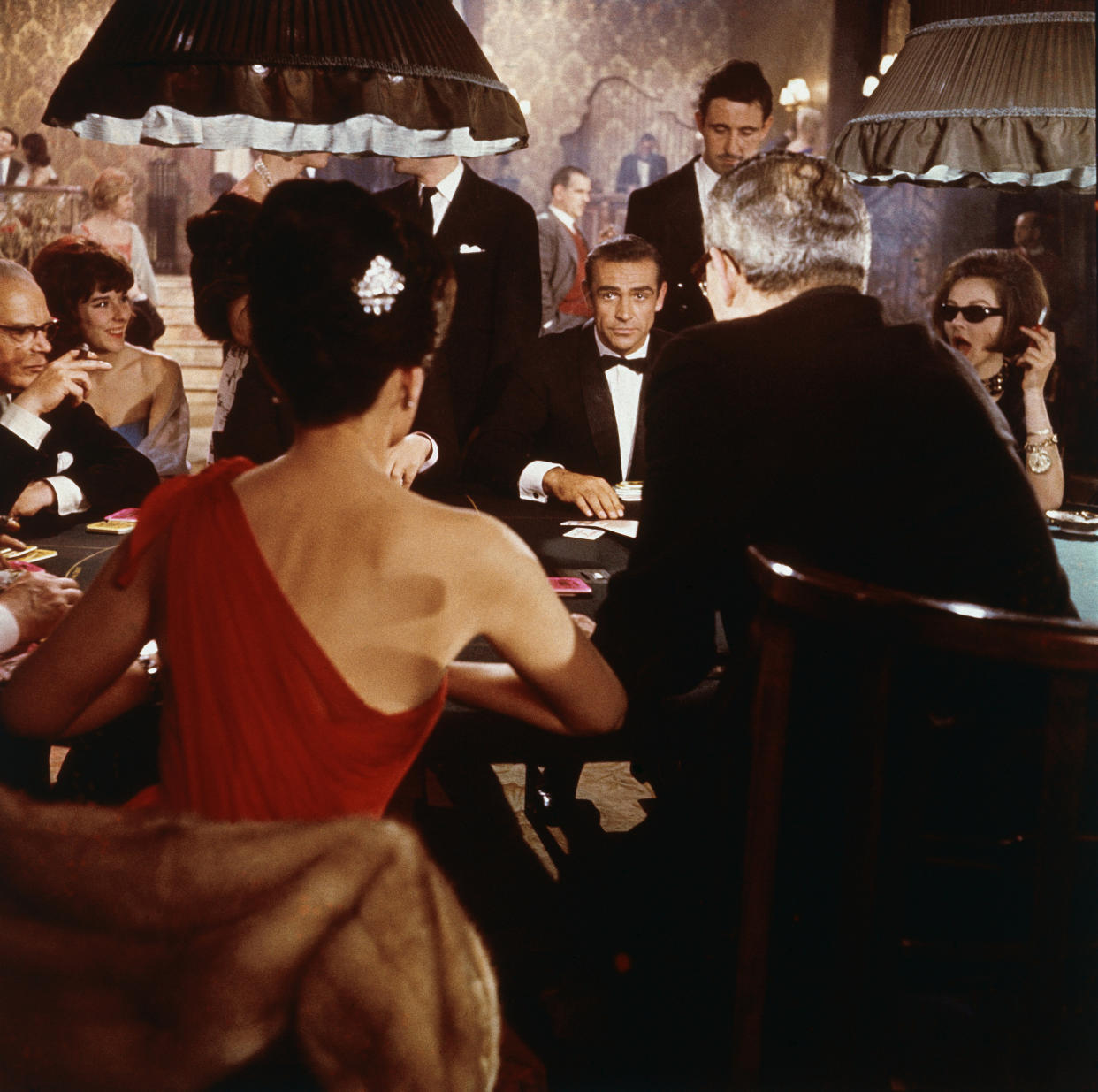 Scottish actor Sean Connery (center) as fictional secret agent James Bond sits at a casino card table in a scene from the film 'Dr. No,' directed by Terence Young, 1962. British actress Eunice Gayson sits with her back to the camera in a red, off the shoulder dress. (Photo by MGM Studios/Courtesy of Getty Images)
