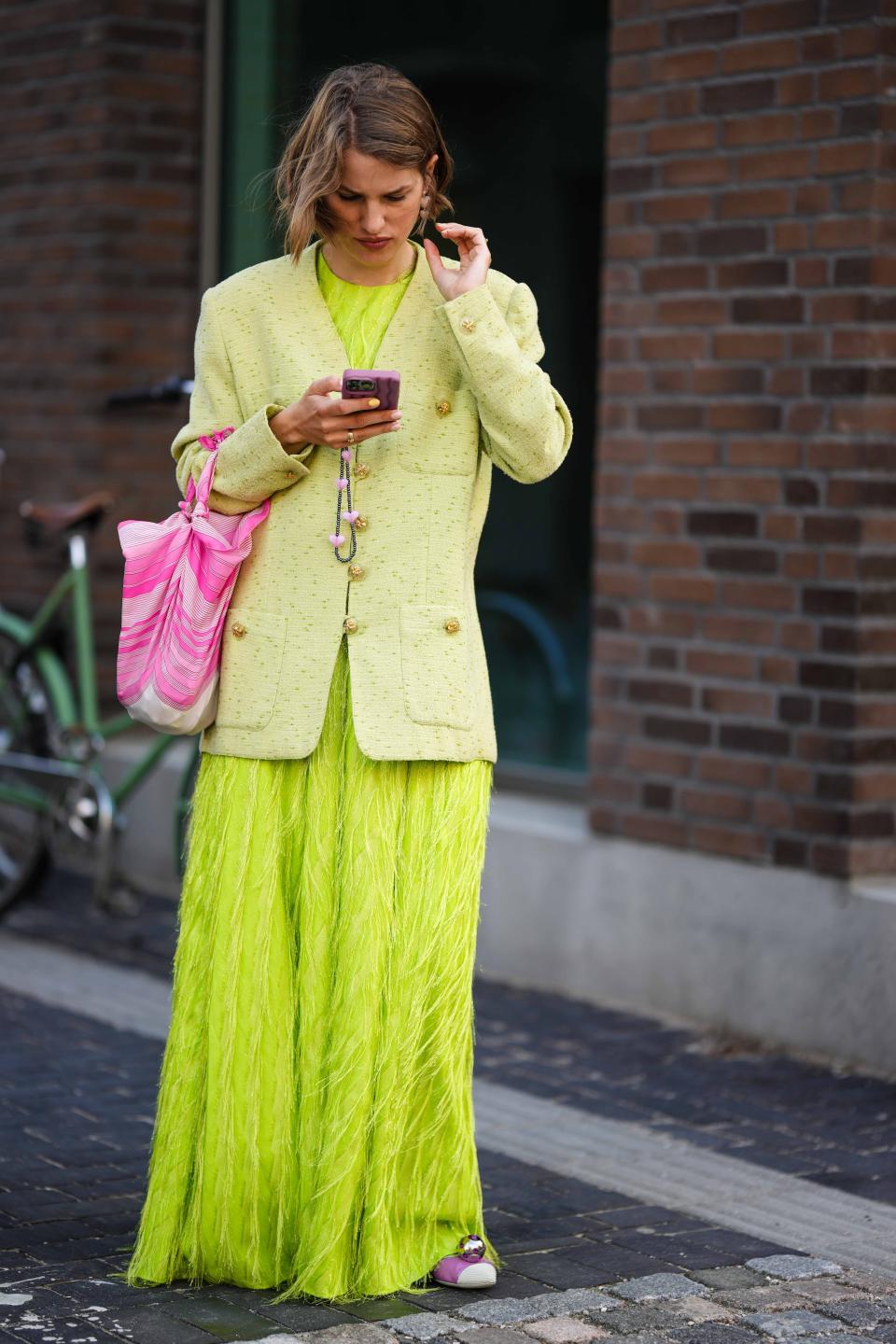 A fashion show attendee sporting a neon dress and blazer scrolls on her phone outside the venue