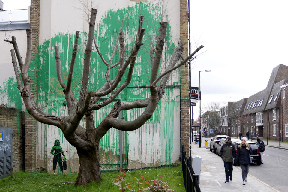 People look at a new Banksy painting on a wall in London, Monday, March 18, 2024. A new Banksy mural drew crowds to a London street on Monday, even before the elusive graffiti artist confirmed that the work was his. The artwork in the Finsbury Park neighborhood covers the wall of a four-story building and shows a small figure holding a pressure hose beside a real tree. Green paint has been sprayed across the wall, replicating the absent leaves of the tree, which has been severely cropped. Banksy claimed the work by posting before and after photos of the location on his official Instagram account.(AP Photo/Alastair Grant)