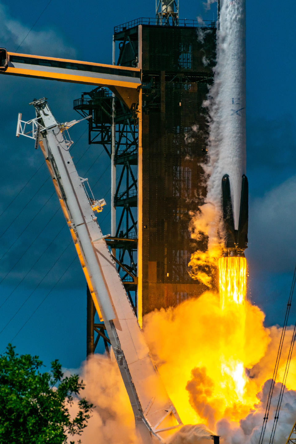 a white rocket with stowed/folded black landing legs is launching. yellow orange fire spews from its engines above a glowing plume. a support tower leans away from the ascending rocket. a launch tower and access arm stand behind. the sky is blue.