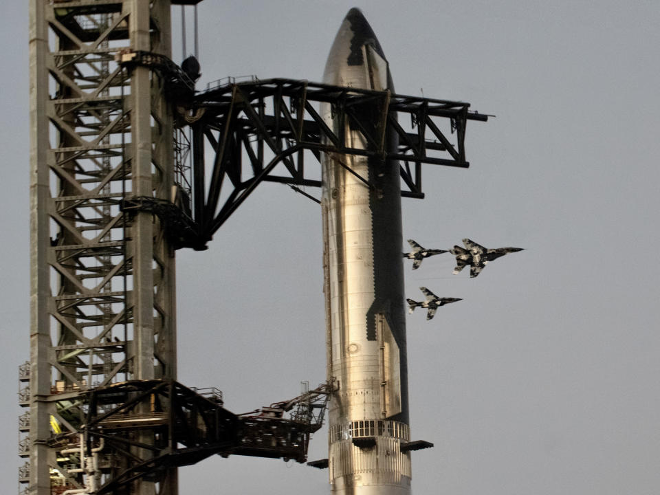 Three white and grey camo jets fly in formation, emerging from behind a giant and tall stainless steel rocket, secured at the top by two black support arms connected to an adjacent launch tower.