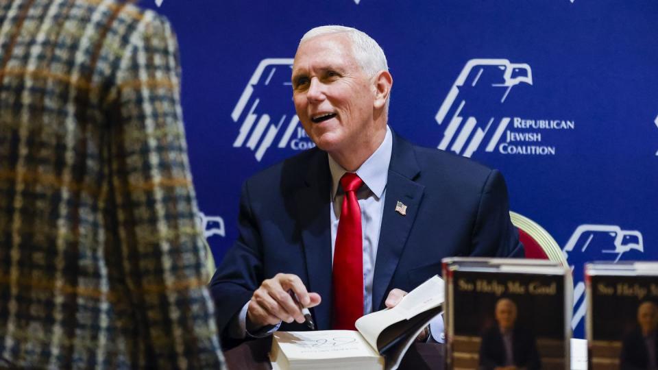mike pence sits at a table and holds open a book to the title page, he looks at someone across from him and smiles, a stack of his book so help me god are just visible in the bottom right corner of the photo