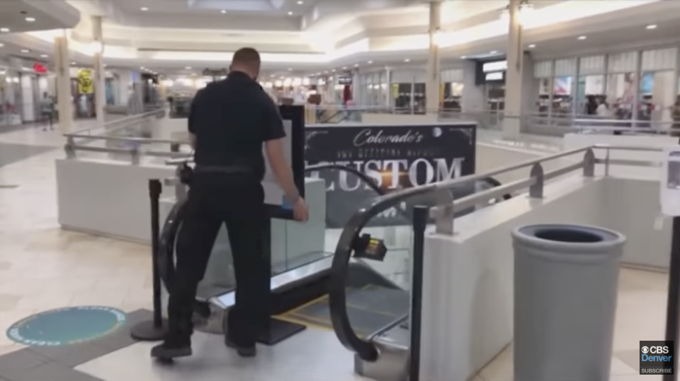 A screenshot from the CBS News Denver story showing a policeman blocking off the top of an escalator in the shopping centre. 