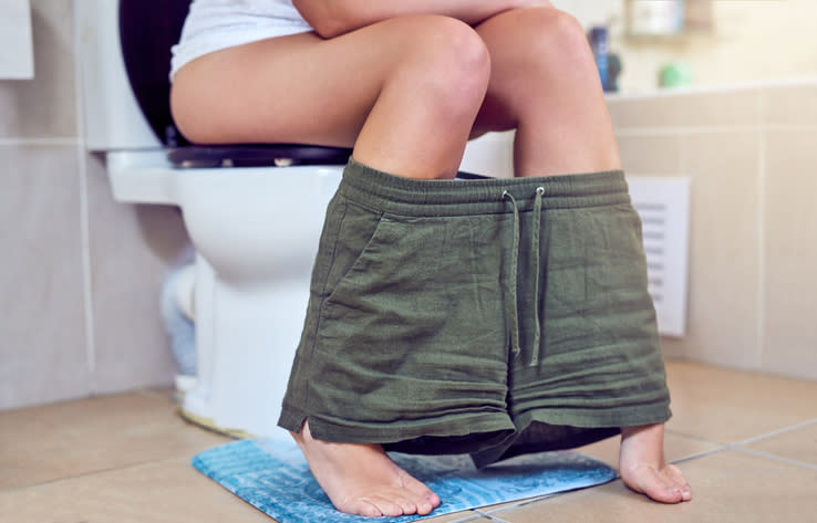 Person seated on a toilet with shorts around their ankles, partial view, focusing on lower body