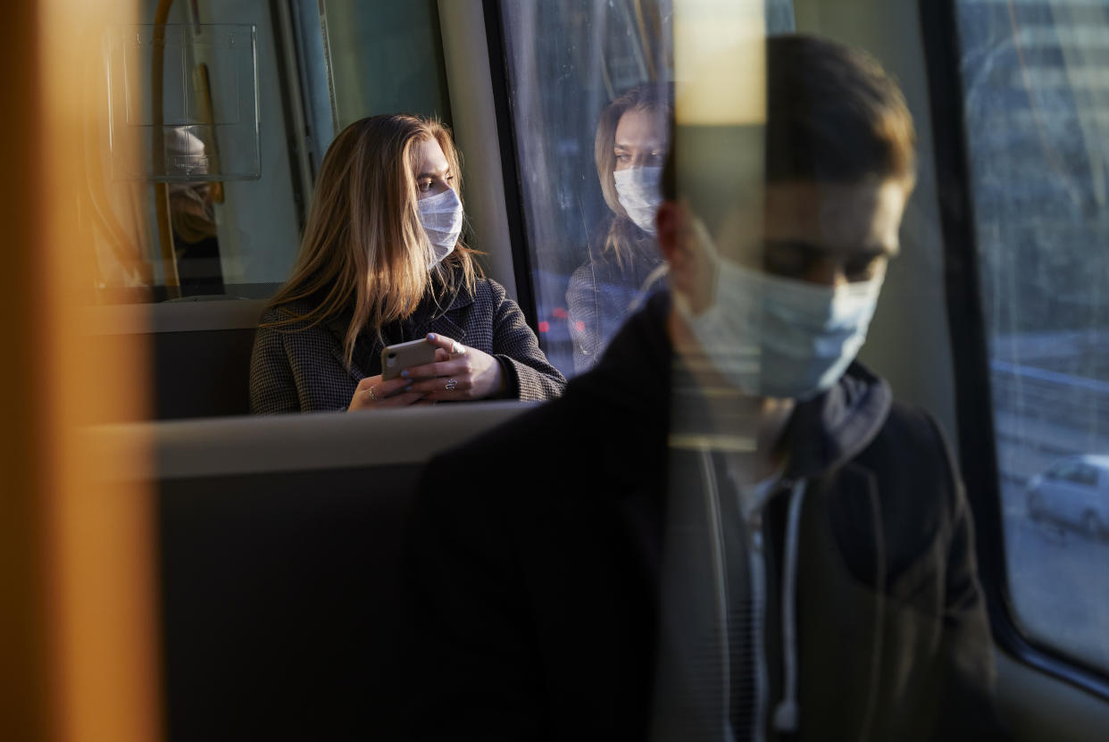 young woman sitting in train wearing protective mask, using smartphone