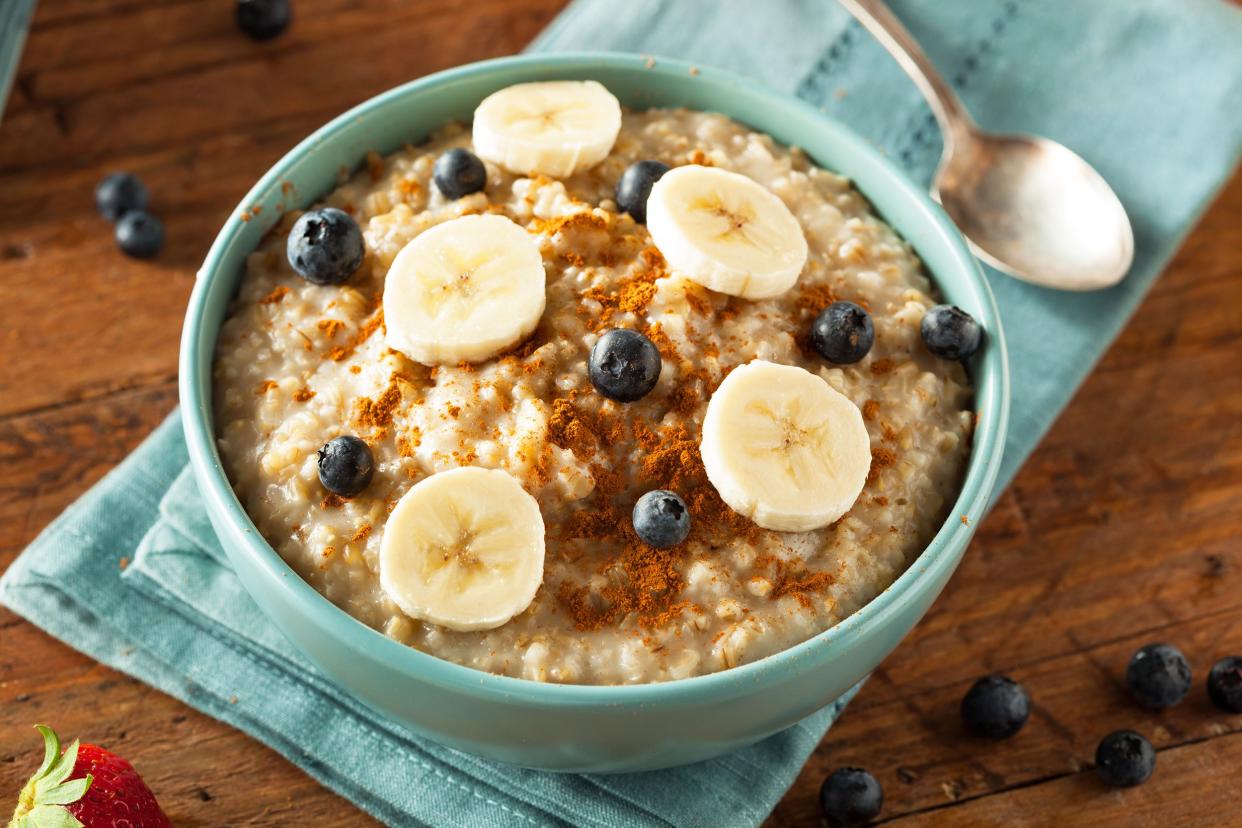 Cooked oatmeal with blueberries and slices of banana in a steel blue bowl, on a steel blue napkin with a spoon, surrounded by blueberries and strawberries on a rustic wooden table