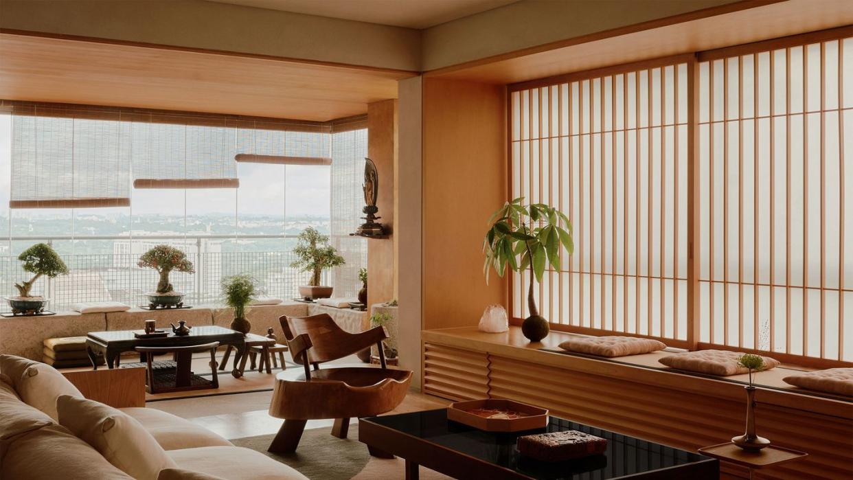 japanese style living room with built in wood storage and window seat with cushions, vertical slats on windows, sofa, cocktail table, chair made from a tree trunk, windows with bamboo blinds, bonsai trees