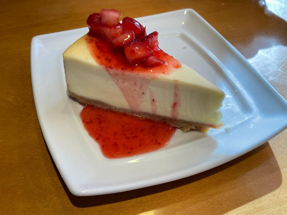 Slice of cheesecake covered in a strawberry sauce and a pile of strawberries on top sits on a square white plate on a brown table at Olive Garden