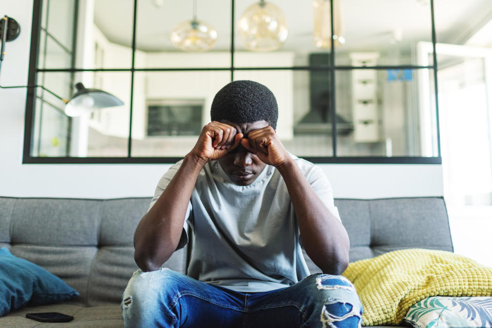 A person sits on a couch in a modern living room, leaning forward with their elbows on their knees and hands clasped in front of their face, appearing distressed